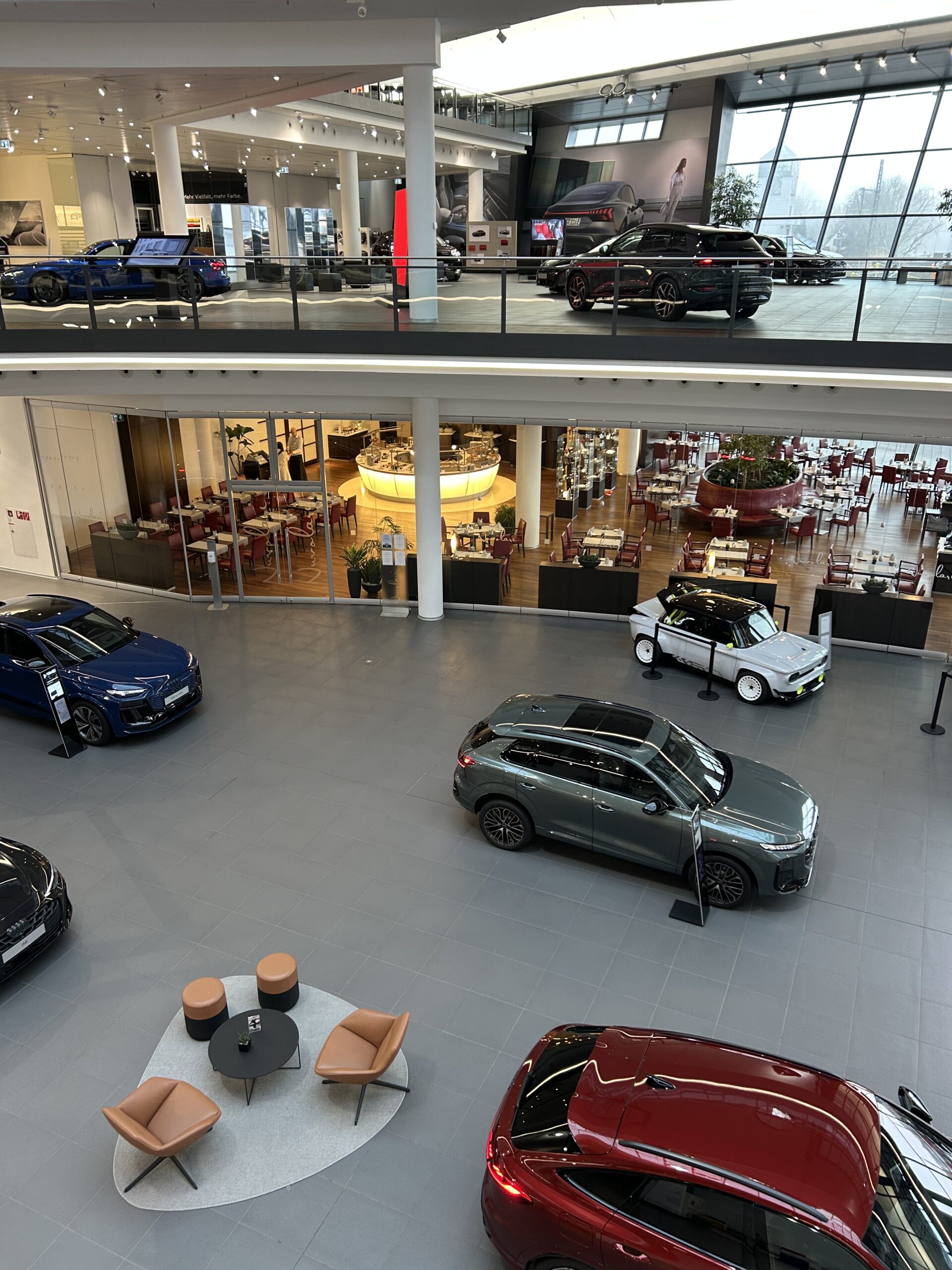 Blick von oben auf die weite Ausstellungshalle mit verschiedenen Audi-Modellen, modernen Sitzgruppen und einem Restaurantbereich im Hintergrund.“