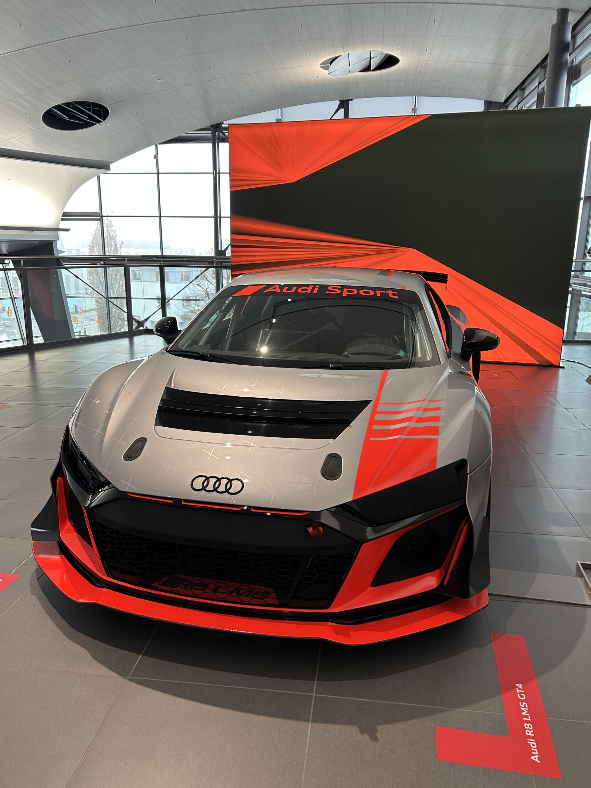 Frontansicht eines silbernen Audi R8 LMS GT4 Rennwagens mit markanten roten Akzenten und 'Audi Sport‘' Schriftzug auf der Windschutzscheibe.