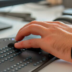 Eine Hand bedient eine Braillezeile vor einem Computerbildschirm – das Gerät übersetzt digitale Inhalte in tastbare Blindenschrift für blinde oder sehbehinderte Menschen.