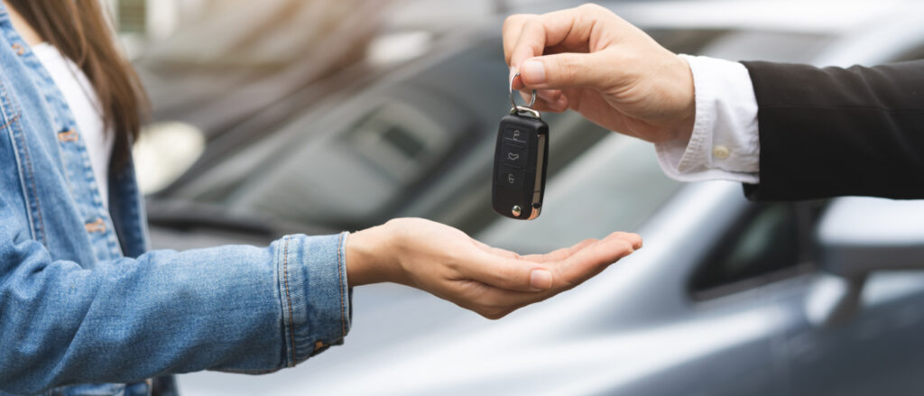 Eine Person übergibt einer anderen Person einen Autoschlüssel von einem im Hintergrund stehendem Auto.
