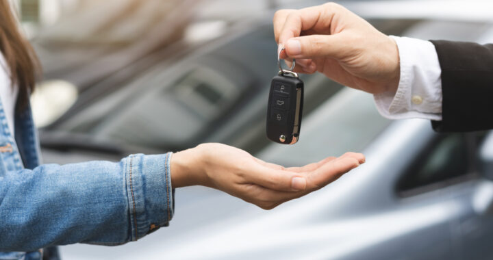Eine Person übergibt einer anderen Person einen Autoschlüssel von einem im Hintergrund stehendem Auto.