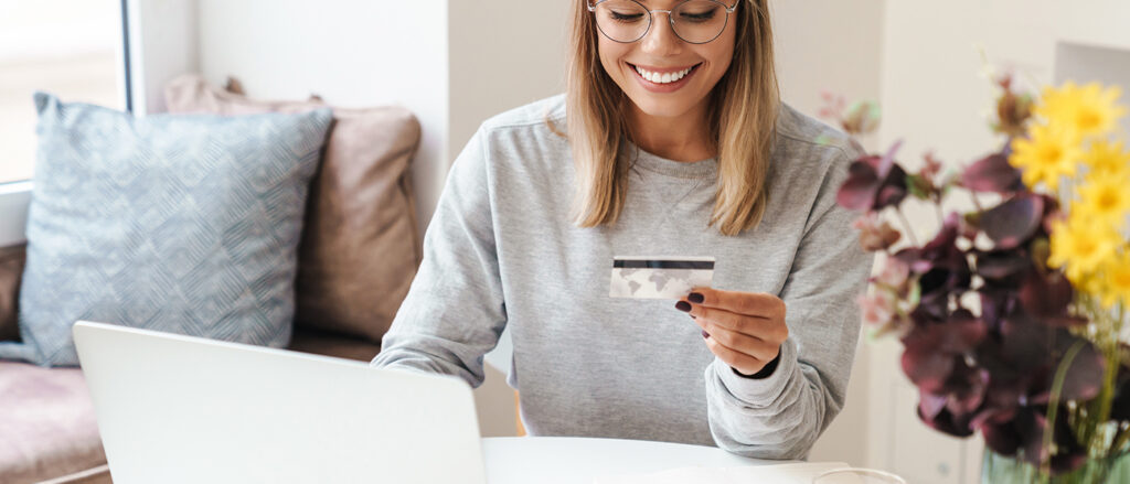 Lächelnde junge Frau mit Brille sitzt am Laptop und hält eine Kreditkarte in der Hand, neben ihr ein Blumenstrauß. Es scheint als kaufe sie grade im Internet ein.