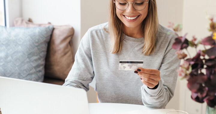 Lächelnde junge Frau mit Brille sitzt am Laptop und hält eine Kreditkarte in der Hand, neben ihr ein Blumenstrauß. Es scheint als kaufe sie grade im Internet ein.