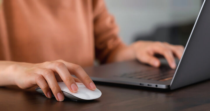 Person bedient mit einer Hand eine Computermaus und mit der anderen ein aufgeklapptes Laptop auf einem Tisch.