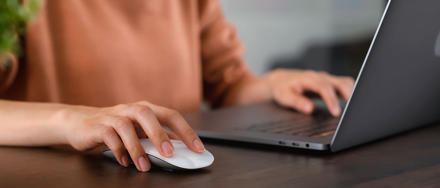 Person bedient mit einer Hand eine Computermaus und mit der anderen ein aufgeklapptes Laptop auf einem Tisch.