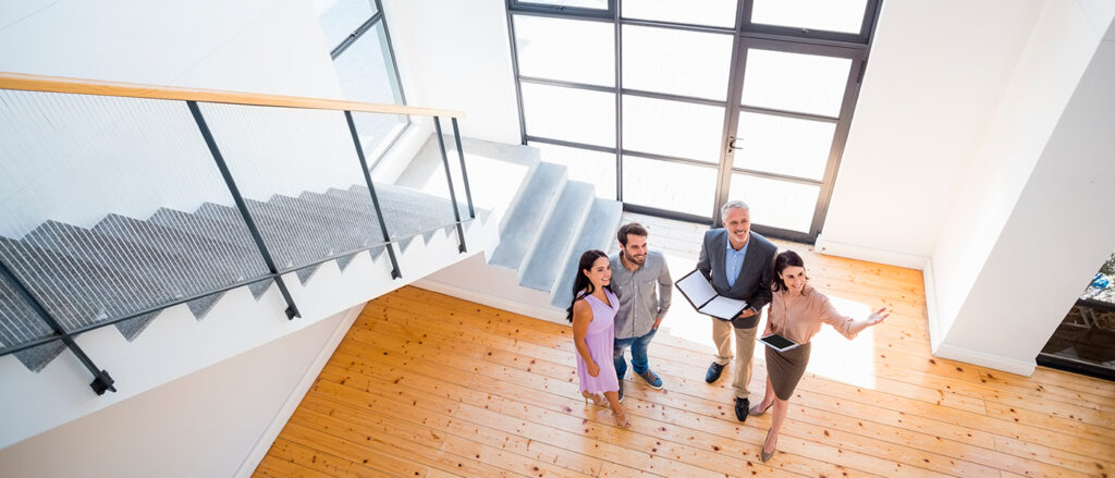 Immobilienmakler zeigen einem Paar ein neues Haus; sie stehen im hohen, lichtdurchfluteten Eingangsbereich.