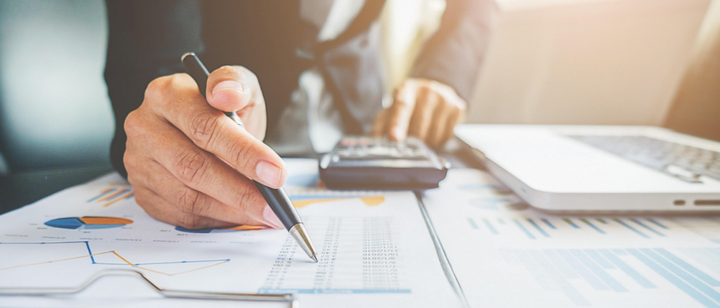 Person in Business-Kleidung hält Stift über einem Blatt mit Statistiken und Tabellen, im Hintergrund sind Taschenrechner und Laptop erkennbar.