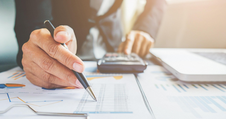 Person in Business-Kleidung hält Stift über einem Blatt mit Statistiken und Tabellen, im Hintergrund sind Taschenrechner und Laptop erkennbar.