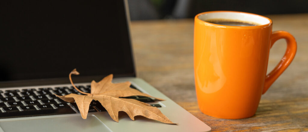 Ein orangefarbener mit Kaffee gefüllter Kaffeebecher steht neben einem Laptop mit einem trockenen Ahornblatt auf der Tastatur.