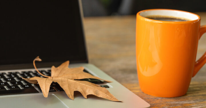 Ein orangefarbener mit Kaffee gefüllter Kaffeebecher steht neben einem Laptop mit einem trockenen Ahornblatt auf der Tastatur.