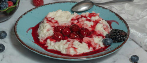 Milchreis auf blauem Teller mit Kirschen und Beeren und einem Löffel am Tellerrand.