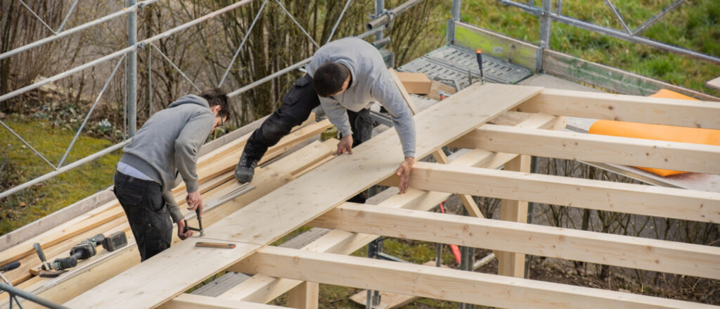 Zwei Handwerker verlegen Holzbretter auf einer Dachkonstruktion aus Holz, zum Bau eines Carports. Sie sind von einem Baugerüst umgeben.