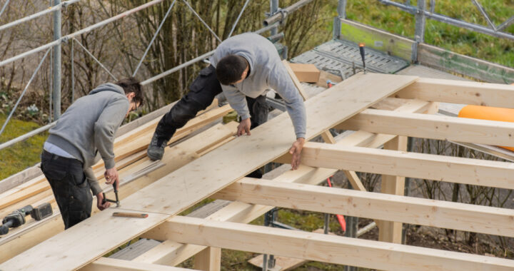 Zwei Handwerker verlegen Holzbretter auf einer Dachkonstruktion aus Holz, zum Bau eines Carports. Sie sind von einem Baugerüst umgeben.