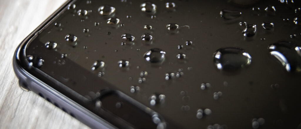 Nahaufnahme eines Ausschnitts eines schwarzen Smartphones mit mehreren Wassertropfen auf dem Bildschirm.