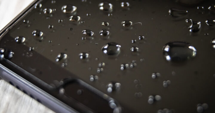 Nahaufnahme eines Ausschnitts eines schwarzen Smartphones mit mehreren Wassertropfen auf dem Bildschirm.