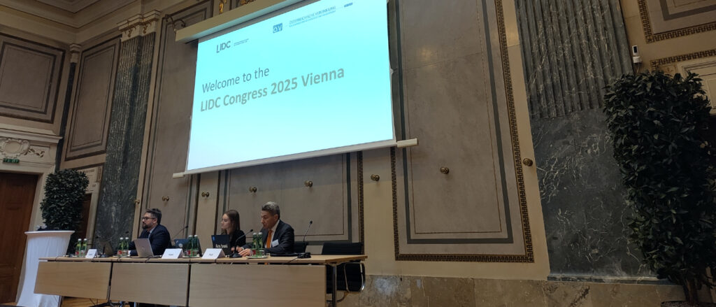Podium mit drei Personen, in ihrem Hintergrund eine große Leinwand auf der eingeblendet ist 'Welcome to the LIDC Congress 2025 Vienna'.