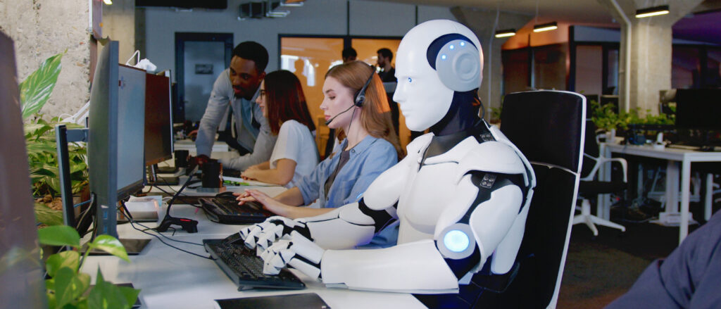Ein humanoider Roboter und eine Frau mit Headset sitzen nebeneinander an Computern in einem modernen Großraumbüro.