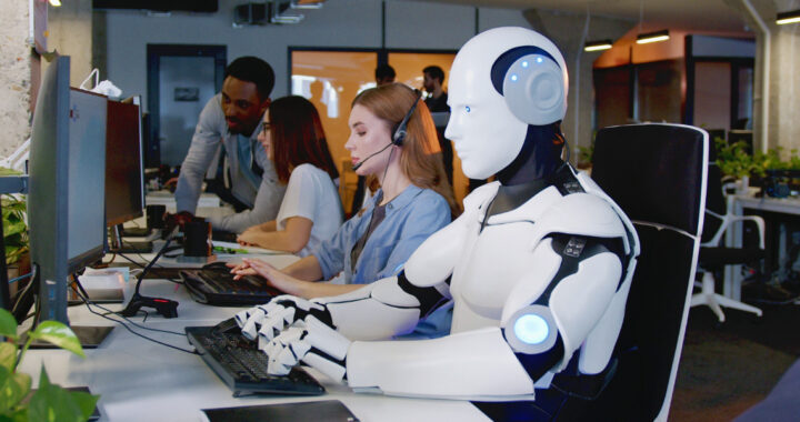 Ein humanoider Roboter und eine Frau mit Headset sitzen nebeneinander an Computern in einem modernen Großraumbüro.