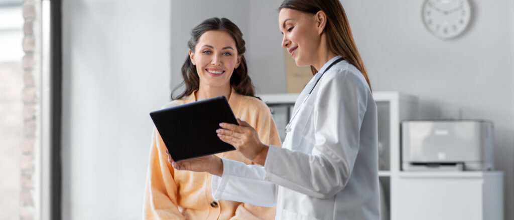 Eine Ärztin zeigt einer lächelnden Patientin ein Tablet in einem hellen Behandlungsraum.