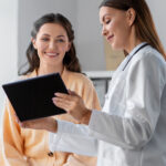 Eine Ärztin zeigt einer lächelnden Patientin ein Tablet in einem hellen Behandlungsraum.