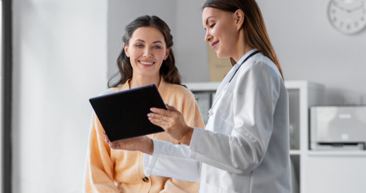 Eine Ärztin zeigt einer lächelnden Patientin ein Tablet in einem hellen Behandlungsraum.