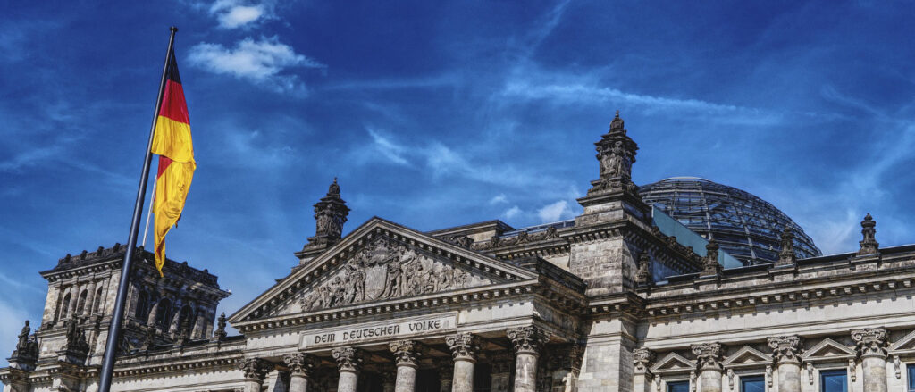 Der Reichstag in Berlin mit gläserner Kuppel und zwei wehenden Deutschlandflaggen bei blauem Himmel.