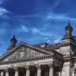Der Reichstag in Berlin mit gläserner Kuppel und zwei wehenden Deutschlandflaggen bei blauem Himmel.