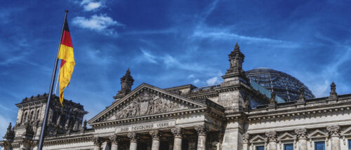 Der Reichstag in Berlin mit gläserner Kuppel und zwei wehenden Deutschlandflaggen bei blauem Himmel.