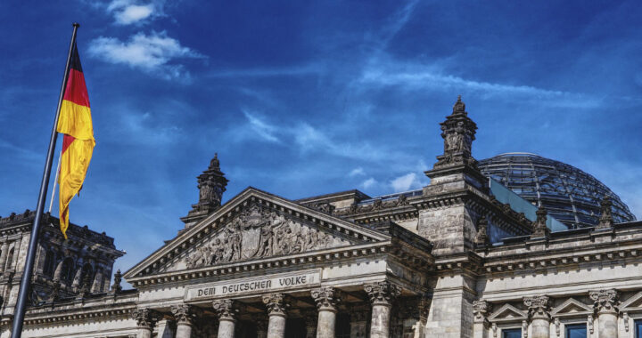Der Reichstag in Berlin mit gläserner Kuppel und zwei wehenden Deutschlandflaggen bei blauem Himmel.