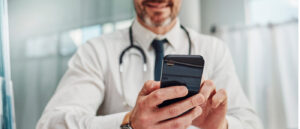 Ein Arzt in weißem Kittel mit Stethoskop um den Hals hält ein Smartphone in beiden Händen und schaut darauf.