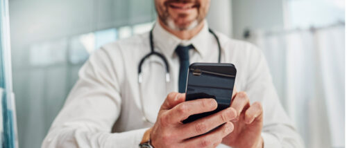 Ein Arzt in weißem Kittel mit Stethoskop um den Hals hält ein Smartphone in beiden Händen und schaut darauf.