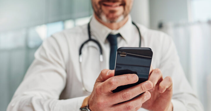 Ein Arzt in weißem Kittel mit Stethoskop um den Hals hält ein Smartphone in beiden Händen und schaut darauf.