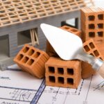 Ein Modellhaus, Ziegelsteine und ein Spachtel stehen/liegen auf einer Bauzeichnung