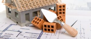 Ein Modellhaus, Ziegelsteine und ein Spachtel stehen/liegen auf einer Bauzeichnung