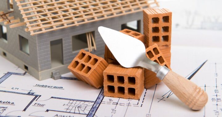 Ein Modellhaus, Ziegelsteine und ein Spachtel stehen/liegen auf einer Bauzeichnung