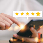 Eine Person hält ein Smartphone in den Händen und tippt auf das Display. Über dem Handy erscheint eine weiße Sprechblase mit fünf goldenen Sternen, die eine positive Bewertung symbolisieren. Der Hintergrund ist hell und leicht unscharf.