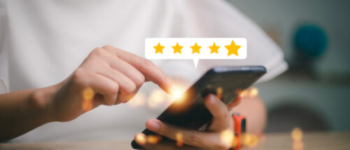 Eine Person hält ein Smartphone in den Händen und tippt auf das Display. Über dem Handy erscheint eine weiße Sprechblase mit fünf goldenen Sternen, die eine positive Bewertung symbolisieren. Der Hintergrund ist hell und leicht unscharf.