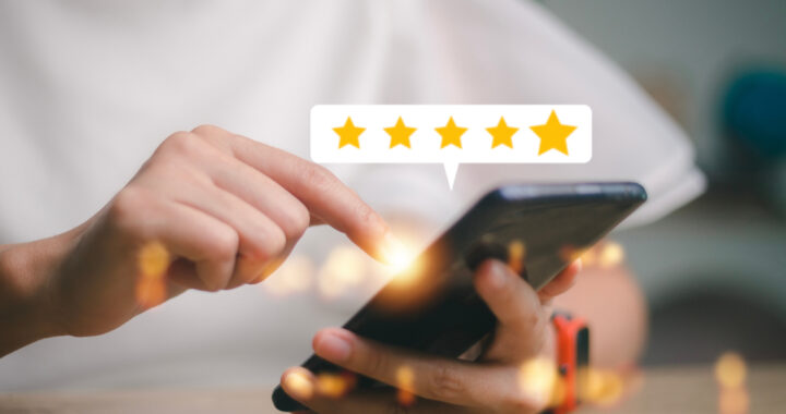 Eine Person hält ein Smartphone in den Händen und tippt auf das Display. Über dem Handy erscheint eine weiße Sprechblase mit fünf goldenen Sternen, die eine positive Bewertung symbolisieren. Der Hintergrund ist hell und leicht unscharf.