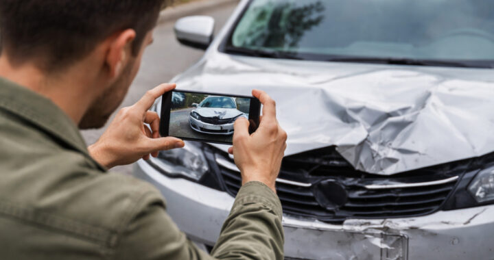 Nahaufnahme einer Person von hinten, die mit einem Smartphone ein Foto von der beschädigten Front eines silbernen Autos macht. Die Motorhaube des Fahrzeugs weist deutliche Unfallspuren auf.