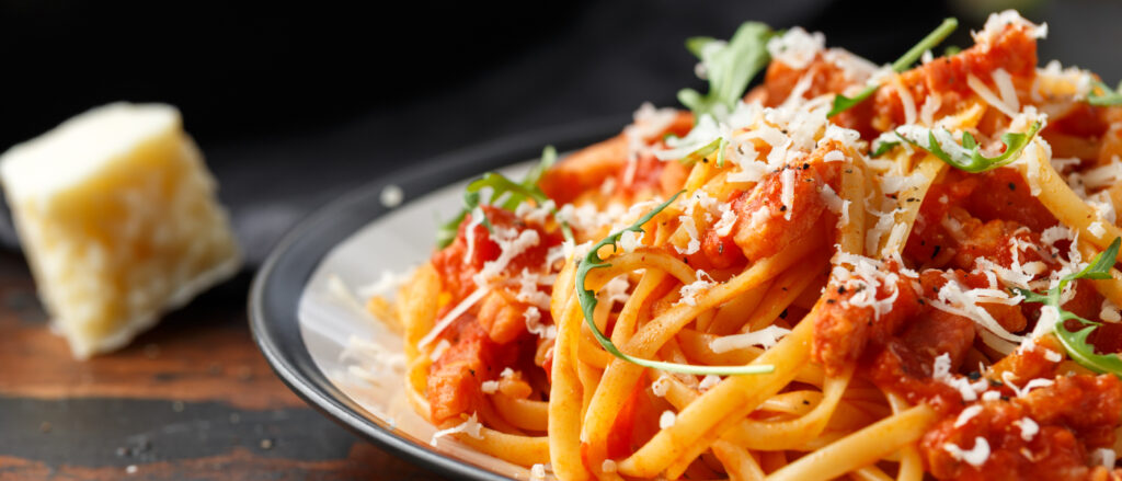 Nahaufnahme eines Tellers Pasta in einer rötlichen Tomaten-Sauce, garniert mit fein geriebenem Hartkäse und frischen grünen Rucola-Blättern. Im unscharfen Hintergrund liegt ein Stück Käse auf einer dunklen, rustikalen Holzoberfläche.
