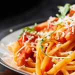Nahaufnahme eines Tellers Pasta in einer rötlichen Tomaten-Sauce, garniert mit fein geriebenem Hartkäse und frischen grünen Rucola-Blättern. Im unscharfen Hintergrund liegt ein Stück Käse auf einer dunklen, rustikalen Holzoberfläche.