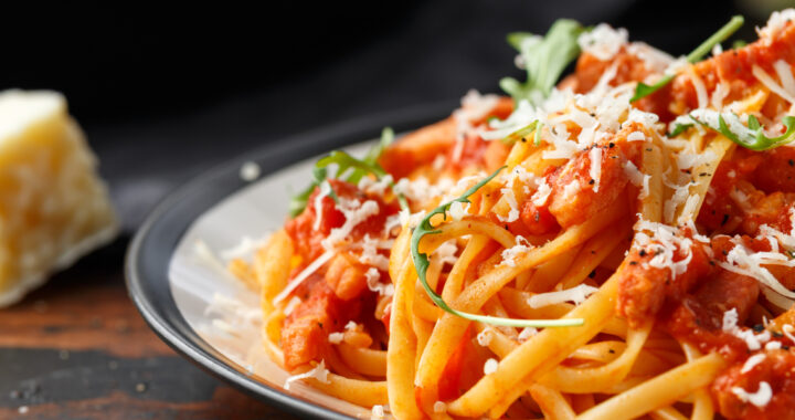 Nahaufnahme eines Tellers Pasta in einer rötlichen Tomaten-Sauce, garniert mit fein geriebenem Hartkäse und frischen grünen Rucola-Blättern. Im unscharfen Hintergrund liegt ein Stück Käse auf einer dunklen, rustikalen Holzoberfläche.