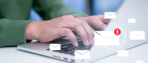 Nahaufnahme von Händen, die auf einer Laptop-Tastatur tippen, während zahlreiche digitale Briefumschläge auf den Bildschirm einströmen. Ein rotes Ausrufezeichen markiert eine Nachricht als Warnung vor unzulässigen Werbemails.