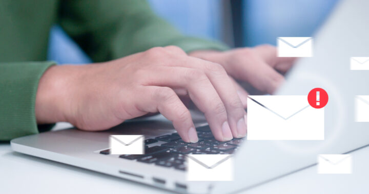 Nahaufnahme von Händen, die auf einer Laptop-Tastatur tippen, während zahlreiche digitale Briefumschläge auf den Bildschirm einströmen. Ein rotes Ausrufezeichen markiert eine Nachricht als Warnung vor unzulässigen Werbemails.