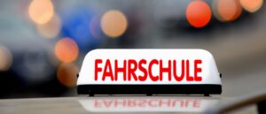 Nahaufnahme eines beleuchteten weißen Fahrschulschilds mit roter Aufschrift „FAHRSCHULE“ auf einem Fahrzeugdach vor unscharfem Lichthintergrund.
