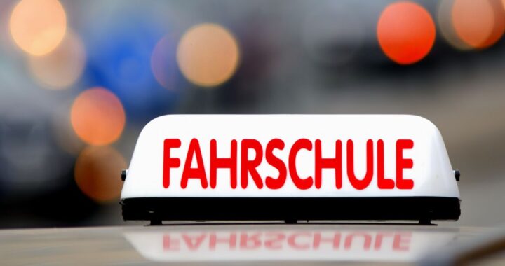 Nahaufnahme eines beleuchteten weißen Fahrschulschilds mit roter Aufschrift „FAHRSCHULE“ auf einem Fahrzeugdach vor unscharfem Lichthintergrund.