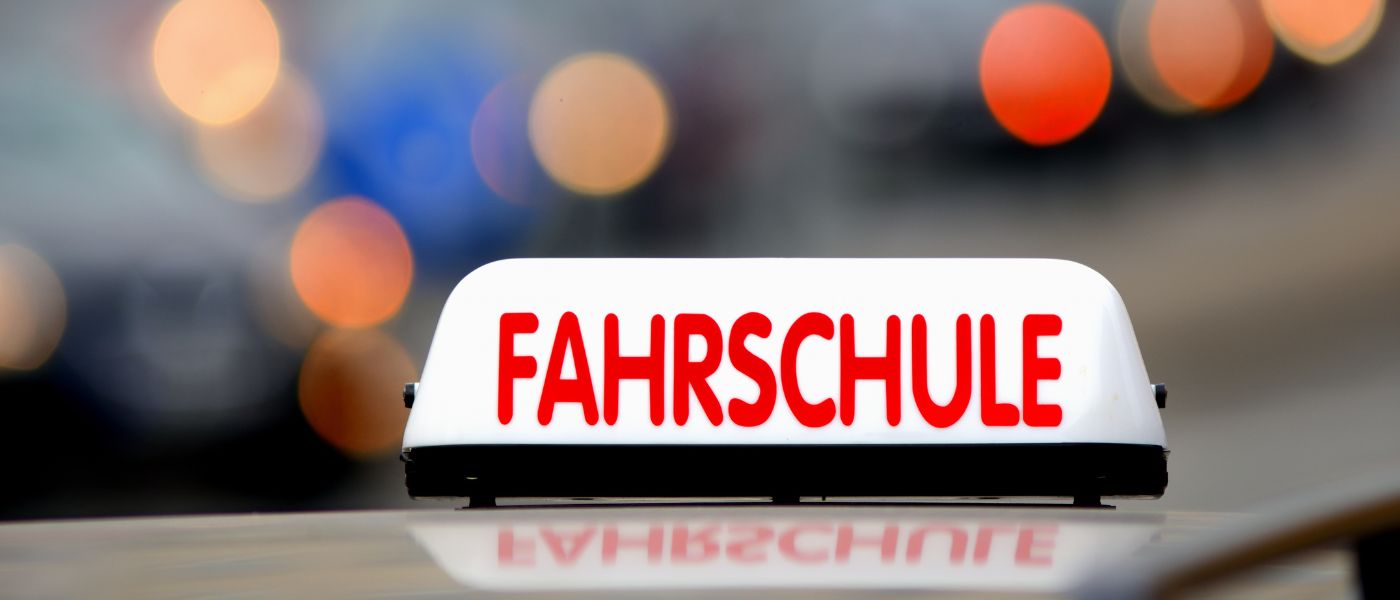 Nahaufnahme eines beleuchteten weißen Fahrschulschilds mit roter Aufschrift „FAHRSCHULE“ auf einem Fahrzeugdach vor unscharfem Lichthintergrund.