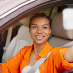 Eine lächelnde junge Automotive-Influencerin sitzt am Steuer eines Autos und macht mit ihrem Smartphone ein Selfie durch das offene Seitenfenster. Sie trägt ein leuchtend orangefarbenes Hemd und ist angeschnallt.