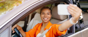 Eine lächelnde junge Automotive-Influencerin sitzt am Steuer eines Autos und macht mit ihrem Smartphone ein Selfie durch das offene Seitenfenster. Sie trägt ein leuchtend orangefarbenes Hemd und ist angeschnallt.