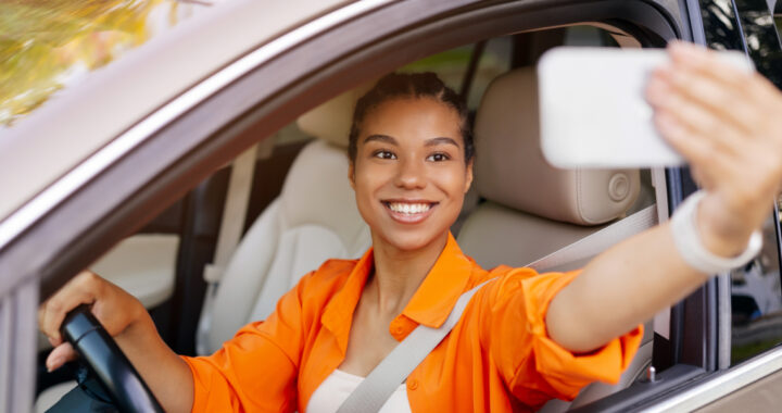 Eine lächelnde junge Automotive-Influencerin sitzt am Steuer eines Autos und macht mit ihrem Smartphone ein Selfie durch das offene Seitenfenster. Sie trägt ein leuchtend orangefarbenes Hemd und ist angeschnallt.
